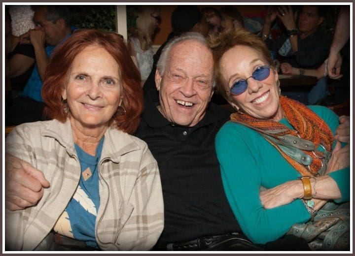 Three People Sitting Side by Side, With a Woman on the Left Wearing a Plaid Jacket Over a Blue Shirt, a Person in the Middle Wearing a Black Shirt, and a Woman on the Right in a Vibrant Green Top With an Orange Scarf.